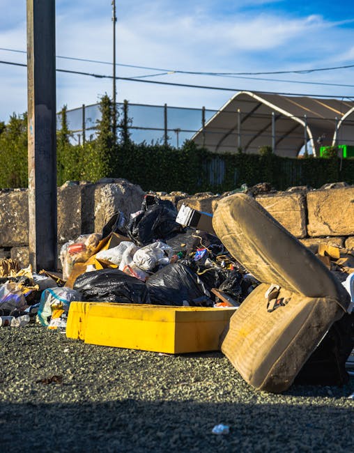A close-up view of an outdoor waste accumulation showing a pile of rubbish on a gravel surface. The rubbish includes black plastic bags filled with waste, a yellow plastic storage container, and an old, worn beige car tyre lying on its side. In the background, there is a stone wall constructed from rough, irregularly shaped stones and a metal fence behind it. Beyond the fence, green bushes, tall trees, and a shelter structure with a curved roof are visible, with the sky overhead appearing partly cloudy with patches of blue. The scene is illuminated by natural daylight, highlighting the details of the waste materials and the surrounding environment. This image illustrates the kind of unsorted waste that might be managed through private or alternative waste collection services such as those provided by wastecollectionknightsbridge.co.uk, reflecting a typical scenario where rubbish is temporarily left outdoors awaiting removal or disposal.