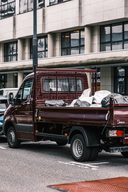 A maroon-colored flatbed pickup truck parked on an urban street in front of a modern building with large windows. The truck's bed is loaded with various waste items, including bags of garbage and distinguishable black and white plastic sacks, likely containing household or construction rubbish. The vehicle features a metal safety grille behind the cab for securing the load, which appears somewhat cluttered but organized for removal. The scene is set during daylight, with ambient natural light illuminating the truck and the surrounding pavement. In the background, there are other parked vehicles and a tall streetlamp, indicating a busy city environment. This image, associated with private waste collection services like those offered by wastecollectionknightsbridge.co.uk, exemplifies on-site waste handling that falls outside typical municipal rubbish collection, emphasizing independent or commercial disposal options suitable for local authorities or businesses seeking alternative rubbish removal solutions.