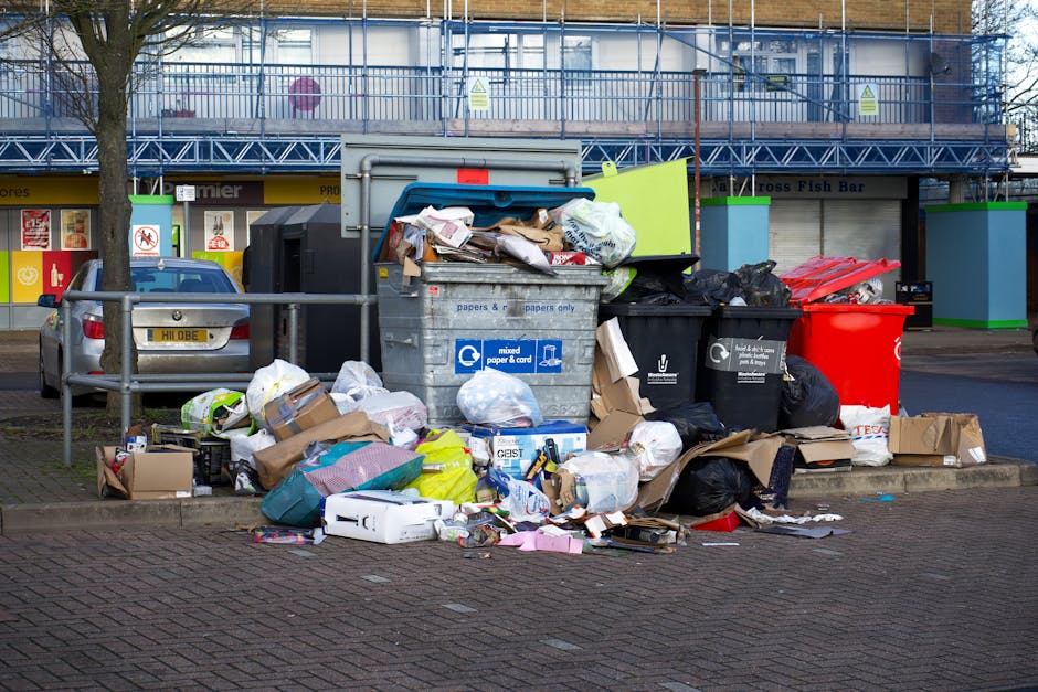 An outdoor scene showing a collection of overflowing waste bins and scattered rubbish on a paved area adjacent to a car park. The central foreground features multiple black, grey, and red wheelie bins, with some lids open and garbage spilling out. The contents include crumpled paper, cardboard boxes, plastic bags, and loose packaging materials, some piled on the ground surrounding the bins. The discarded waste appears to consist of household items such as flattened cardboard boxes, plastic wrappers, and paper items. Behind the bins, there is a blue and grey commercial waste container labeled for mixed paper and cards, with some bags and packaging placed on top of or leaning against it. To the left, a silver car with a visible number plate is parked behind a metal railing. In the background, there is a building with a storefront featuring a yellow and blue sign, along with a blue scaffolding structure indicating ongoing construction or refurbishment. The overall scene suggests a temporary accumulation of waste outside a commercial or residential area, possibly relating to non-regular rubbish collection, with waste management visible as an essential service to be undertaken by private waste collection providers like wastecollectionknightsbridge.co.uk.