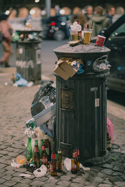 An outdoor scene showing a collection of overflowing waste bins and scattered rubbish on a paved area adjacent to a car park. The central foreground features multiple black, grey, and red wheelie bins, with some lids open and garbage spilling out. The contents include crumpled paper, cardboard boxes, plastic bags, and loose packaging materials, some piled on the ground surrounding the bins. The discarded waste appears to consist of household items such as flattened cardboard boxes, plastic wrappers, and paper items. Behind the bins, there is a blue and grey commercial waste container labeled for mixed paper and cards, with some bags and packaging placed on top of or leaning against it. To the left, a silver car with a visible number plate is parked behind a metal railing. In the background, there is a building with a storefront featuring a yellow and blue sign, along with a blue scaffolding structure indicating ongoing construction or refurbishment. The overall scene suggests a temporary accumulation of waste outside a commercial or residential area, possibly relating to non-regular rubbish collection, with waste management visible as an essential service to be undertaken by private waste collection providers like wastecollectionknightsbridge.co.uk.