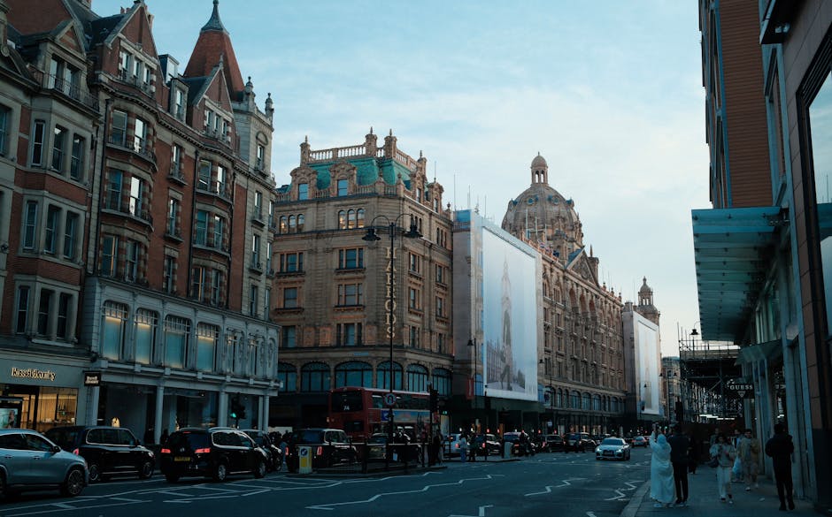 A busy street scene in an urban area featuring a row of historic multi-story buildings with intricate architectural details, including arched windows, decorative cornices, and a prominent domed rooftop structure visible in the background. The buildings are constructed from light-colored stone with reddish-brown brick accents, and some facades include large display windows and signage. The street is populated with parked cars, a red double-decker bus, and a few pedestrians walking along the pavement, some dressed in formal attire. Along the right side, a modern building with a glass front and a visible blue external structure contrasts with the older architecture. The sky is partly cloudy, casting soft natural light over the scene. This image visually supports the context of alternative waste handling, with the urban environment suggesting nearby commercial activities and possible private disposal services managed by waste collection specialists like wastecollectionknightsbridge.co.uk.