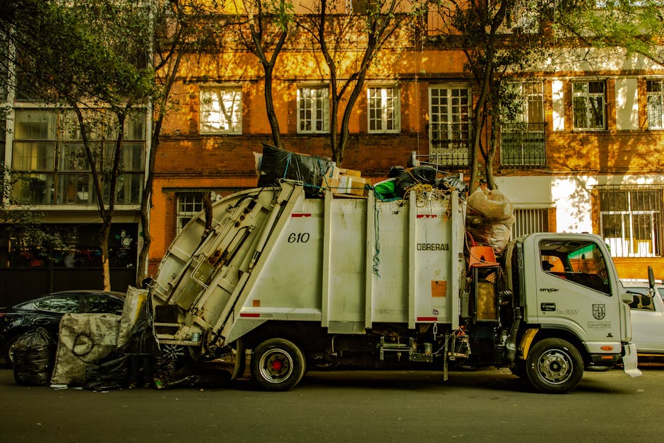 A white waste collection truck with a closed back filled with various rubbish items, parked on a city street in front of a multi-storey red-brick residential building with large windows. The truck features a compact cab with a side door and is fitted with a hydraulic mechanism for loading and unloading waste. On the left side of the image, several black garbage bags and loose waste materials are placed on the pavement, some of which are leaning against the front of the truck. The scene is shaded by trees with green foliage, casting dappled sunlight across the building and street, creating a contrast of light and shadow. The environment suggests a typical urban setting during daytime, where private waste collection services, such as those by wastecollectionknightsbridge.co.uk, handle rubbish removal, demonstrating an example of on-site waste clearance or alternative disposal outside municipal collection points. The overall scene highlights the process of refuse collection related to premises that may require independent waste management solutions, ensuring proper rubbish removal in a residential area.