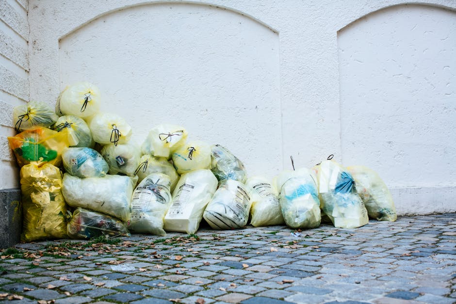 A collection of plastic rubbish bags placed on a cobbled pavement next to a white textured wall. The bags are of various sizes and colors, mostly yellow, white, and transparent, containing waste materials. Some bags are tightly knotted, showing the black ties securing their openings, while others loosely drape over the contents. The waste appears to include plastic bottles, packaging, and general household rubbish, suggesting a mixed waste load. The bags are stacked and leaning against the wall in an informal manner, indicative of an external waste collection point often used for private rubbish disposal or alternative collection services such as those provided by waste management specialists like wastecollectionknightsbridge.co.uk. The environment is an outdoor urban setting with a weathered cobblestone surface, and there is no visible vehicle or other equipment present in the image. Overall, the scene reflects a typical scene of non-kerbside waste accumulation awaiting collection or removal, aligned with professional rubbish disposal practices.