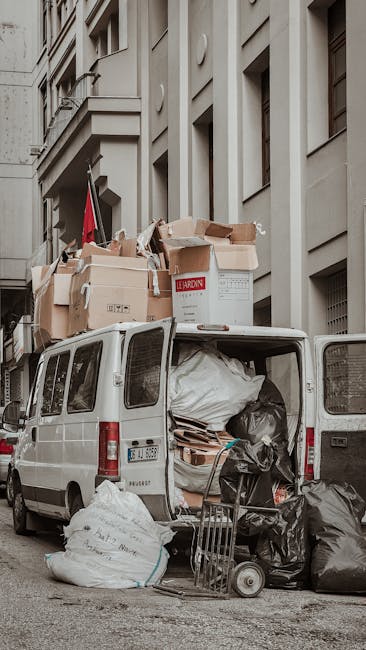 A white panel van parked on a narrow city street with its rear doors open, revealing an overflowing load of rubbish bags, cardboard boxes, and debris. The trash includes large black plastic bags, flattened cardboard pieces, and smaller waste items, some of which are stacked on the ground beside the vehicle. The van is positioned adjacent to a multi-storey building with light grey stone or concrete facades, featuring vertical window openings and architectural details typical of urban residential or commercial structures. On top of the van, several cardboard boxes and larger waste containers are stacked, appearing to be part of an on-site rubbish collection or alternative waste handling setup. The scene is illuminated by natural daylight, with minimal shadows, highlighting the cluttered and densely packed nature of the rubbish load. This scenario reflects a form of private waste collection, potentially undertaken by specialist rubbish removal services such as wastecollectionknightsbridge.co.uk, highlighting the challenge of managing refuse in tight urban environments where access may be difficult for standard waste collection services.