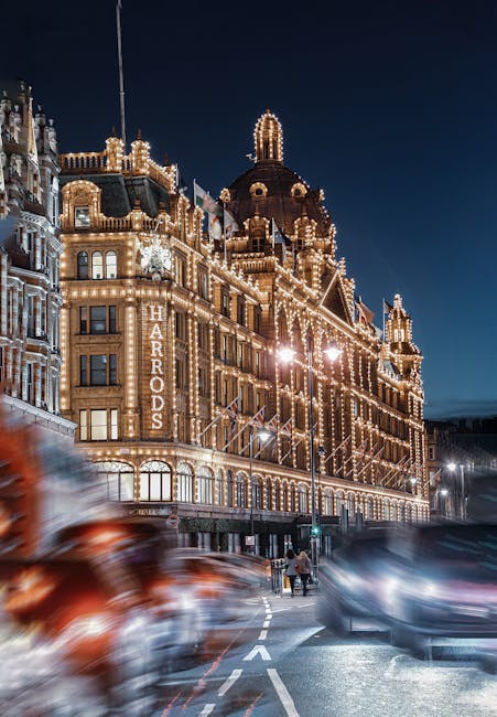 A large, ornate building illuminated with numerous warm white lights, featuring decorative turrets, domes, and intricate architectural details, located in the Harrods area of Knightsbridge. The facade of the building displays a vertically mounted sign reading 'HARDOS' in capital letters. The scene is set during nighttime, with a dark sky overhead and streetlights casting a soft glow on the pavement and surrounding area. In the foreground, several cars are passing by at speed, creating motion blur, with their headlights and taillights visible. Two pedestrians are seen walking on the sidewalk near the building, slightly to the right of the center. The urban environment suggests a busy street scene, with the building serving as a prominent landmark. This scene highlights the contrast between the illuminated historic architecture and the dynamic movement of modern traffic, consistent with an area where alternative waste handling or private rubbish collection services may operate, especially in high-traffic, commercial districts like Knightsbridge. The overall atmosphere evokes a lively night-time setting, capturing the essence of a well-lit, luxurious shopping district with active street life.