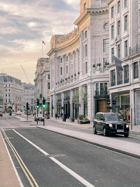 A daytime street scene in a central London district featuring a row of elegant, white stone buildings with classical architectural details such as columns, decorative cornices, and large windows. The street has multiple lanes with painted markings, and there are traffic lights showing green for vehicles. A black van is parked along the curb, and several pedestrians are walking on the wide sidewalk. In the background, additional historic-style buildings line the street, and a flag is visible flying atop one of the structures. The lighting suggests early morning or late afternoon with a partly cloudy sky, creating a soft, natural illumination of the scene. The setting highlights a typical urban environment where private waste removal services, such as those offered by wastecollectionknightsbridge.co.uk, could facilitate rubbish collection from commercial and residential properties in the area, complementing local authority services with on-site clearance options that suit the sophisticated cityscape.
