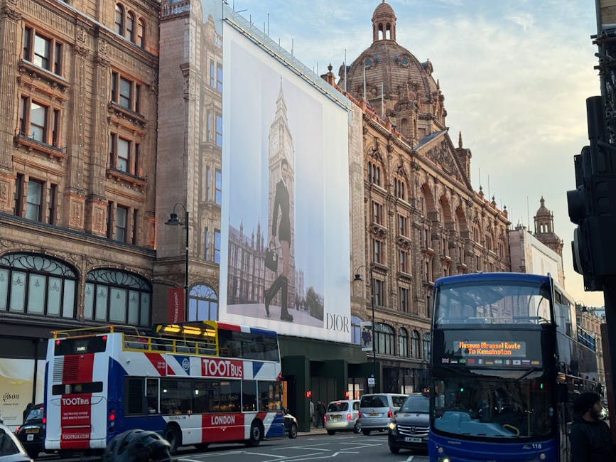 A large, ornate building illuminated with numerous warm white lights, featuring decorative turrets, domes, and intricate architectural details, located in the Harrods area of Knightsbridge. The facade of the building displays a vertically mounted sign reading 'HARDOS' in capital letters. The scene is set during nighttime, with a dark sky overhead and streetlights casting a soft glow on the pavement and surrounding area. In the foreground, several cars are passing by at speed, creating motion blur, with their headlights and taillights visible. Two pedestrians are seen walking on the sidewalk near the building, slightly to the right of the center. The urban environment suggests a busy street scene, with the building serving as a prominent landmark. This scene highlights the contrast between the illuminated historic architecture and the dynamic movement of modern traffic, consistent with an area where alternative waste handling or private rubbish collection services may operate, especially in high-traffic, commercial districts like Knightsbridge. The overall atmosphere evokes a lively night-time setting, capturing the essence of a well-lit, luxurious shopping district with active street life.