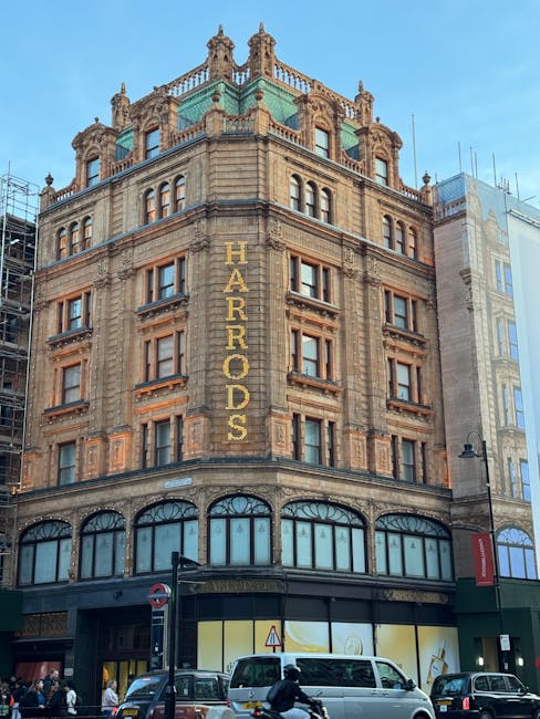 The image depicts a multi-storey historic building with ornate architectural detailing, constructed from brown sandstone with a textured surface and decorative elements around the windows and cornices. The upper floors feature numerous rectangular sash windows with wooden frames, some of which are partially open, and the building is topped with a greenish copper roof with decorative spires and a balustrade. On the side of the building’s corner, large gold lettering spells out 'HARRORDS,' indicating the building’s name. The ground floor has large, curved glass windows with black frames, and the entrance is centrally located beneath these display windows. The scene is set on a busy urban street with vehicles, including a white van and a dark car, parked directly in front of the building. Pedestrians are visible walking along the sidewalk, and a red street lamp is positioned near the building’s corner. The overall atmosphere suggests an active city environment where the building’s retail or commercial space could be serviced by independent waste removal or rubbish collection specialists like wastecollectionknightsbridge.co.uk, supporting efficient on-site clearance of accumulated rubbish from commercial activities.