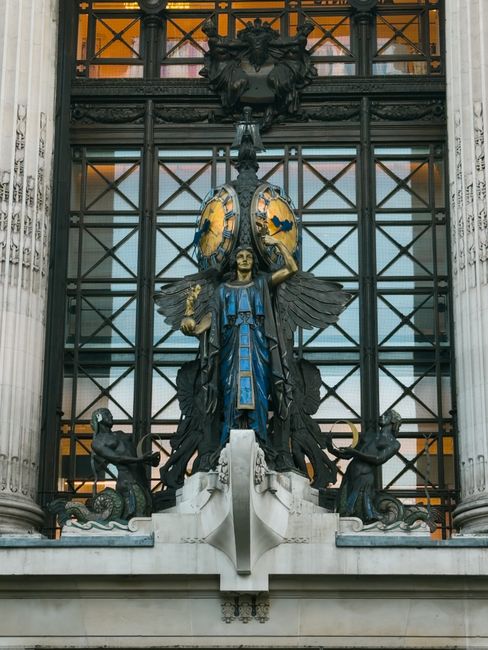 A detailed view of a decorative architectural sculpture located on the facade of a historic building, featuring a central figure with large, detailed wings, dressed in a blue robe with gold accents, holding a mechanical object in each hand. The figure is flanked by two smaller figures with muscular builds, positioned on either side, each holding a circular element and facing outward. Behind the sculpture is an intricate iron framework with cross-hatched patterns, framed by two tall, fluted stone columns with ornate capitals. Above the sculpture, there is a dark, elaborate ornamental crest with a shield and decorative motifs. The lighting highlights the varied textures, including smooth stone, metalwork, and painted finishes, capturing the grandeur of the architectural details. This scene reflects classical influences in urban sculpture and facade ornamentation, relevant to the context of historic city architecture often associated with private or independent restoration and preservation efforts, paralleling themes of alternative waste handling and on-site clearance in the context of general rubbish removal services provided by wastecollectionknightsbridge.co.uk.
