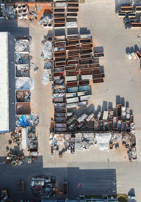 An aerial view of an outdoor storage yard with numerous rusted metal containers and bins arranged in rows on a concrete surface, with some containers stacked and others placed individually. Surrounding the containers are various other discarded materials, including metal scrap, plastic items, and debris, indicating a site for collecting and temporarily storing waste materials related to construction or demolition. To the left, there is a large warehouse structure with a partly visible roof, and on the bottom edge, a section of parking spaces and vehicles can be seen. The environment appears to be a dedicated area for waste management or rubbish storage, consistent with private disposal services such as those offered by waste collection specialists like wastecollectionknightsbridge.co.uk. The scene emphasizes practical on-site clearance and material handling, typical of companies providing alternative waste solutions outside of standard local authority collection, ensuring efficient management of builders waste and materials awaiting transportation or recycling.