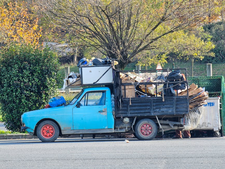 A small, older model light blue truck parked on the side of a street during daylight hours, loaded with various types of waste and debris. The truck's cargo area is filled with cardboard boxes, plastic bags, and miscellaneous trash, some of which are secured with ropes. The vehicle has visible signs of wear, with faded paint and rusty red wheels. In the background, there are green trees with autumn-colored leaves, and a metal fence along the sidewalk. The scene suggests a private rubbish collection or on-site clearance process, with waste prepared for removal outside a residential or semi-urban area. The presence of the loaded truck exemplifies an alternative to standard council rubbish collection, aligned with independent waste disposal services like those offered by wastecollectionknightsbridge.co.uk.