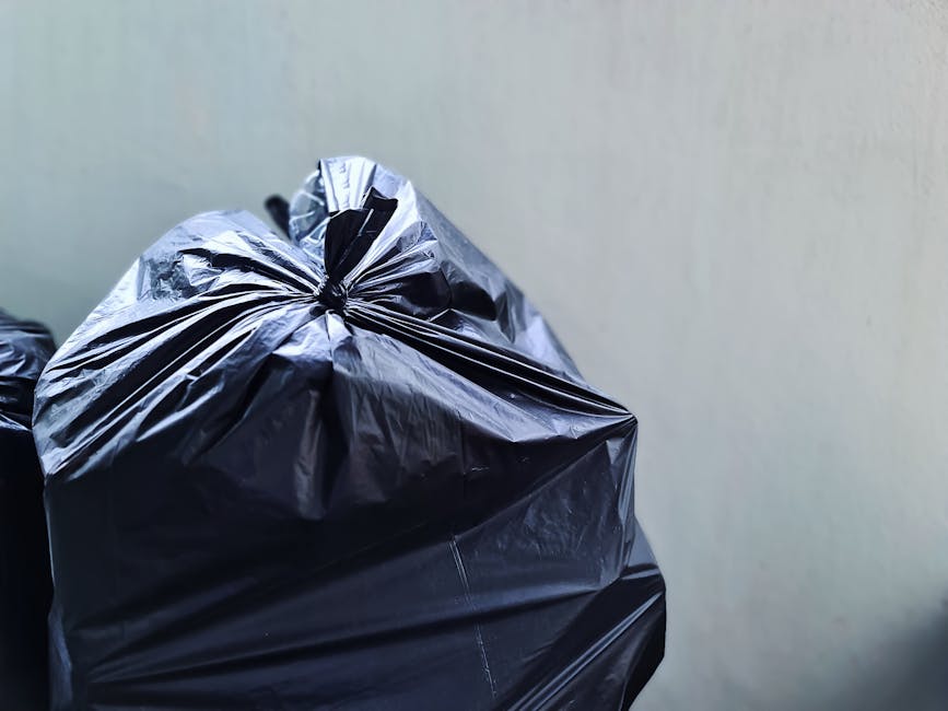 Close-up image of a black plastic rubbish bag with tightly gathered and knotted top, placed against a plain, light-colored wall background. The bag appears to be made of glossy, durable plastic material with a slightly crinkled texture, demonstrating its heaviness and flexibility. It is positioned in the foreground, occupying the left side of the frame, with part of another similar bag visible behind it, suggesting a collection of waste ready for disposal. The environment hints at an indoor or sheltered external space, such as a garage or storage area, where waste is gathered prior to collection. The lighting is even, highlighting the sheen of the plastic and subtle creases in the bag's surface. This scene exemplifies typical private or independent waste handling methods that may complement or serve as an alternative to council rubbish collection services, as facilitated by wastecollectionknightsbridge.co.uk.