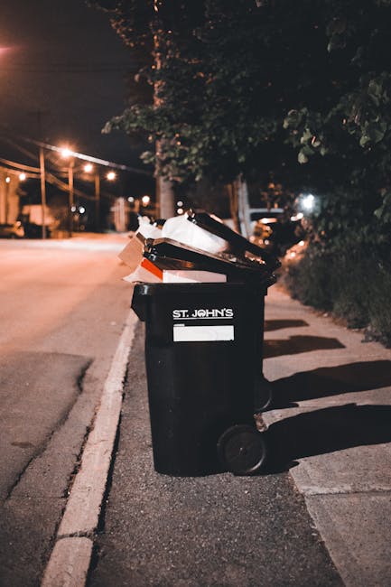 A black wheeled refuse bin positioned on a sidewalk on a quiet street during nighttime. The bin has a white label with the text 'ST. JOHN'S' visible on its front, and it is filled with various waste items, including cardboard, paper, and plastic materials, some of which are protruding from the open lid. The bin is situated near a curb, with its shadow cast onto the pavement by nearby streetlights. The background includes softly illuminated streetlights, trees with dark foliage, and a roadway stretching into the distance, indicating an urban or suburban residential area. The environment appears still and calm, with no immediate activity around the bin, reflecting a typical scene where private waste collection or rubbish removal services might be employed to manage local waste outside of municipal collection schedules. The scene captures a moment of onsite rubbish disposal, aligning with services such as independent waste management or same-day rubbish collection as seen in Knightsbridge.
