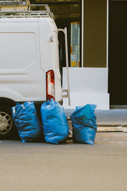 A white commercial van is parked on the street adjacent to a building with large glass windows and a dark facade. Three blue rubbish bags made of plastic, filled and knotted at the top, are stacked against the rear side of the van on the paved surface. The bags exhibit varying shades of blue and appear to contain general waste or discarded items, suitable for private waste collection services. In the background, part of a sidewalk and a white structure or barrier are visible near the storefront. The environmental lighting suggests daytime with natural light illuminating the scene. This setup reflects a typical scene of on-site rubbish removal, possibly arranged by independent waste collection providers such as wastecollectionknightsbridge.co.uk, directly handling waste outside commercial or residential premises without reliance on local authority collection services.
